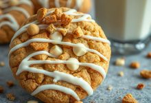 A close-up of a freshly baked cookie topped with white chocolate chips, drizzled with white icing, and sprinkled with crunchy Biscoff cookie pieces. A glass of milk is blurred in the background, complementing the cookie's golden texture and rich toppings.