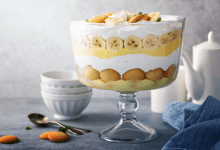 A full view of a glass bowl featuring traditional Southern banana pudding layers: vanilla wafers, fresh banana slices, creamy pudding, and whipped cream, elegantly presented on a table with dessert utensils.