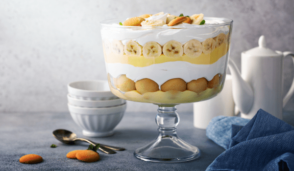 A full view of a glass bowl featuring traditional Southern banana pudding layers: vanilla wafers, fresh banana slices, creamy pudding, and whipped cream, elegantly presented on a table with dessert utensils.