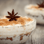 Mexican arroz con leche served in a glass bowl, decorated with a star anise and a cinnamon stick, showcasing layers of texture and color on a light wooden surface.