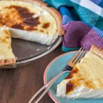 Old Fashioned Custard Pie in a glass pie dish with a golden-brown crust and rich custard filling. A slice is plated on a blue and orange plate, ready to be enjoyed with whipped cream or as is.