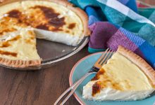 Old Fashioned Custard Pie in a glass pie dish with a golden-brown crust and rich custard filling. A slice is plated on a blue and orange plate, ready to be enjoyed with whipped cream or as is.